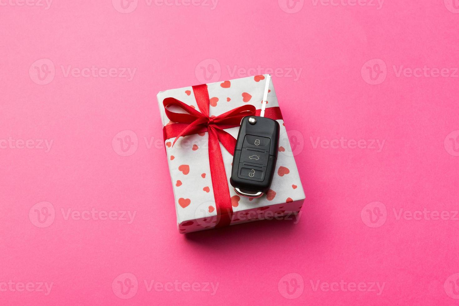 Car key on paper gift box with red ribbon bow and heart on pink table background. Holidays present top view concept photo