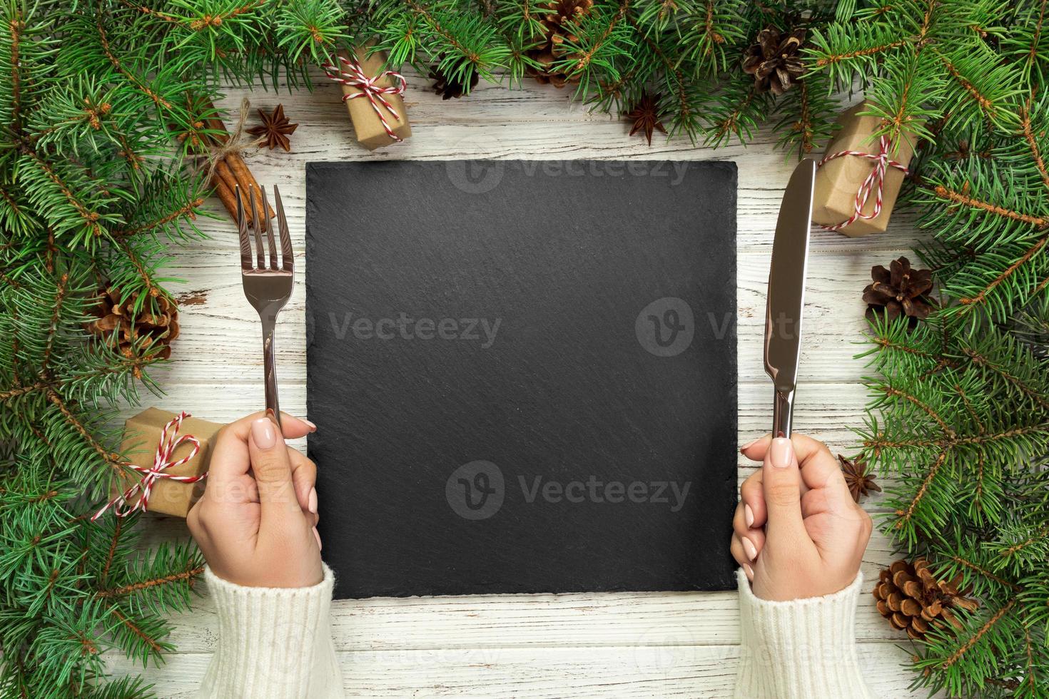 top view girl holds fork and knife in hand and is ready to eat. Empty black slate square plate on wooden christmas background. holiday dinner dish concept with new year decor photo