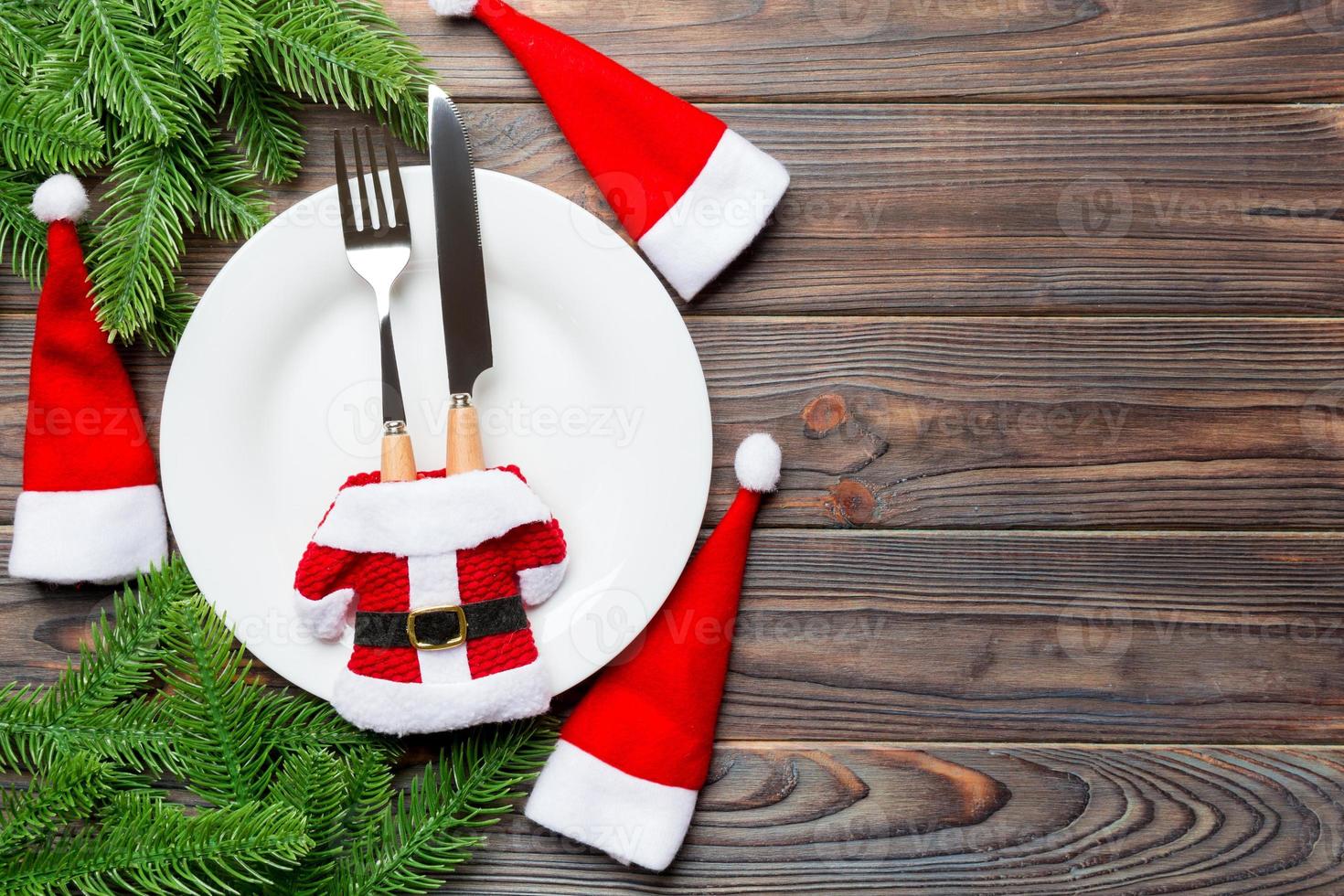 Holiday composition of plate and flatware decorated with Santa hat and clothes on wooden background. Top view of Christmas decorations with empty space for your design. Festive time concept photo