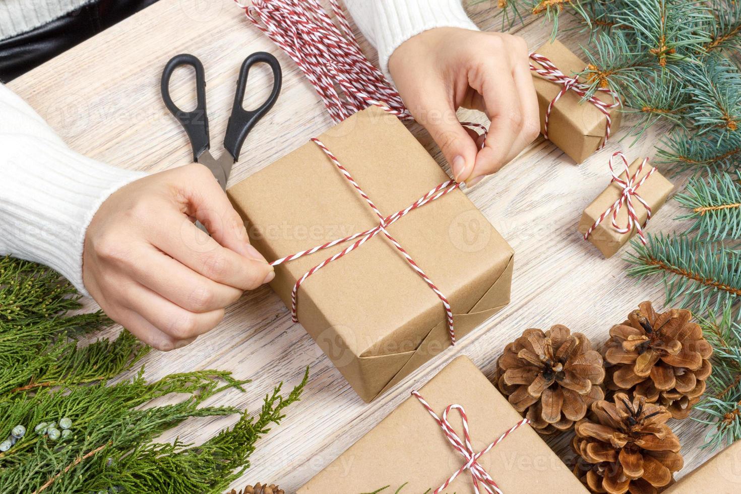 Christmas presents preparation. Gift box wrapped in black and white striped paper, a crate full of pine cones and christmas toys and wrapping materials on a white wood old background photo