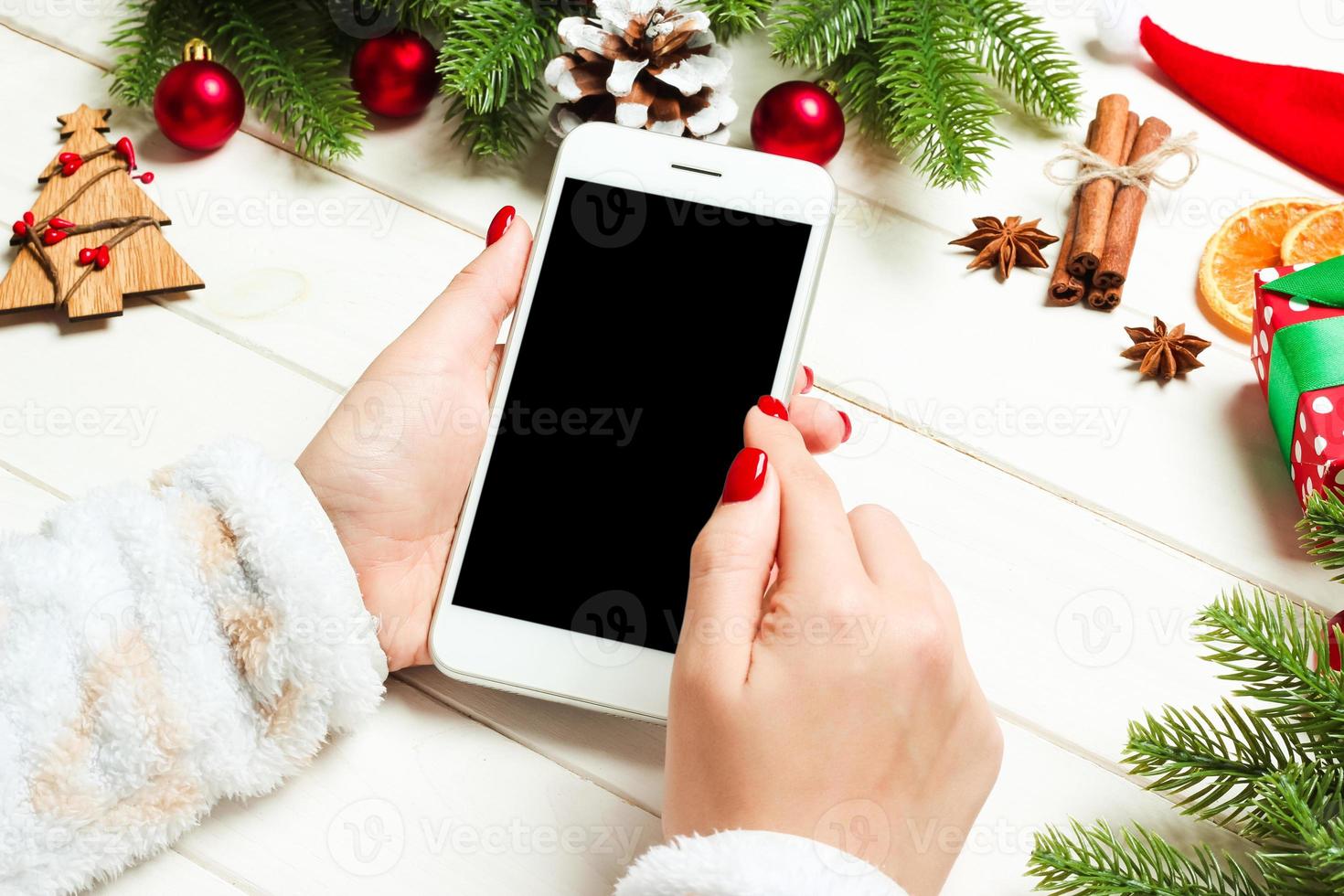 Top view of a woman holding a phone in her hand on wooden New Year background made of fir tree and festive decorations. Christmas holiday concept. Mockup photo