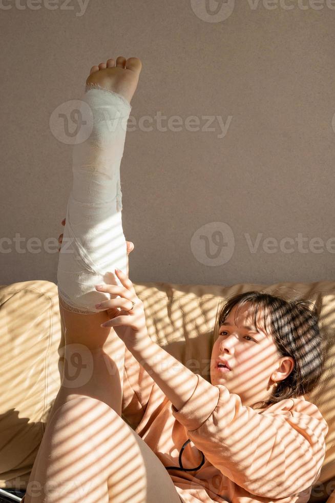 girl with an expression of pain on her face, lies on the couch with her leg raised up in a cast. The girl corrects the plaster on her leg or doing exercises photo