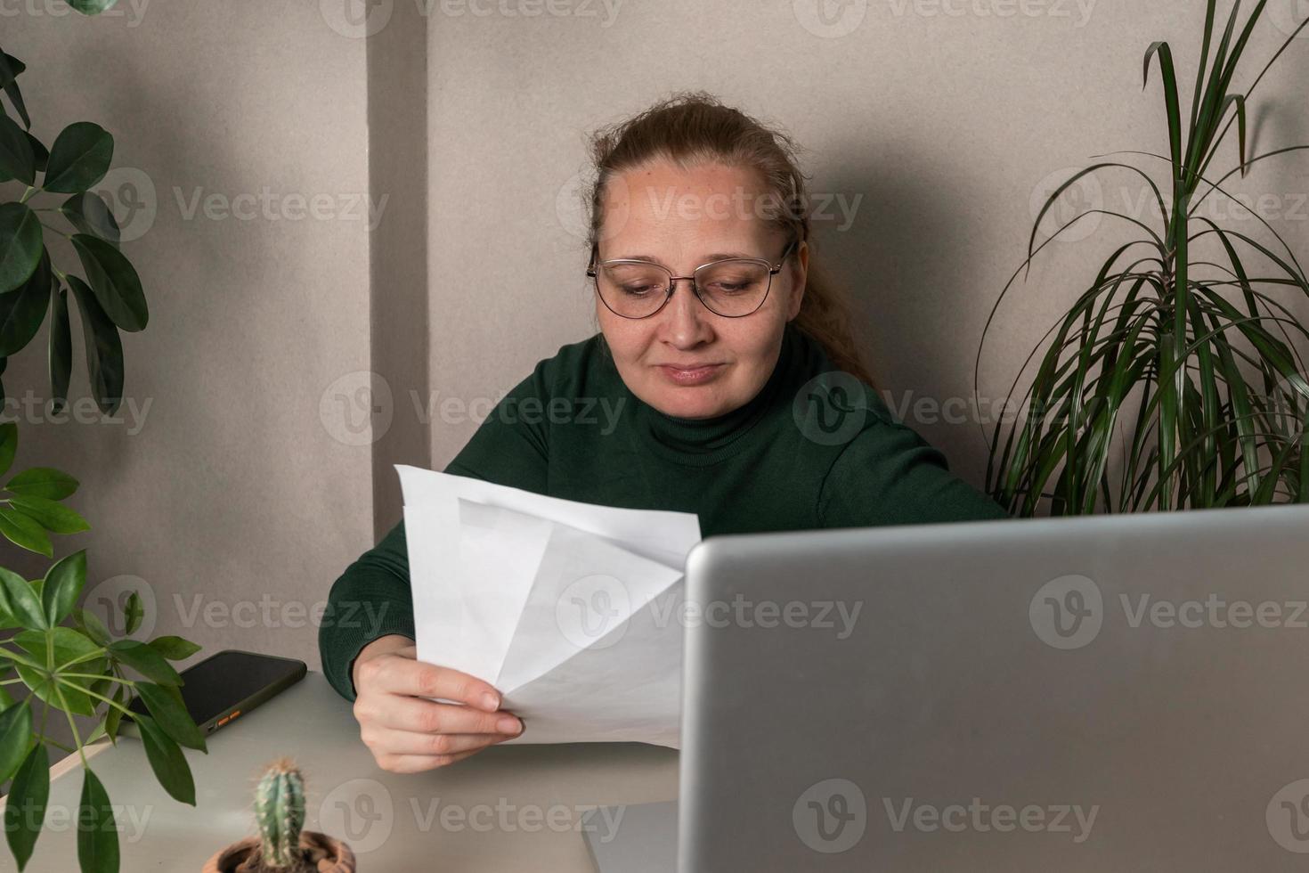 A middle-age businesswoman works at a desk, makes notes in a laptop, and checks the data on her phone. Business, online business, work in the office photo