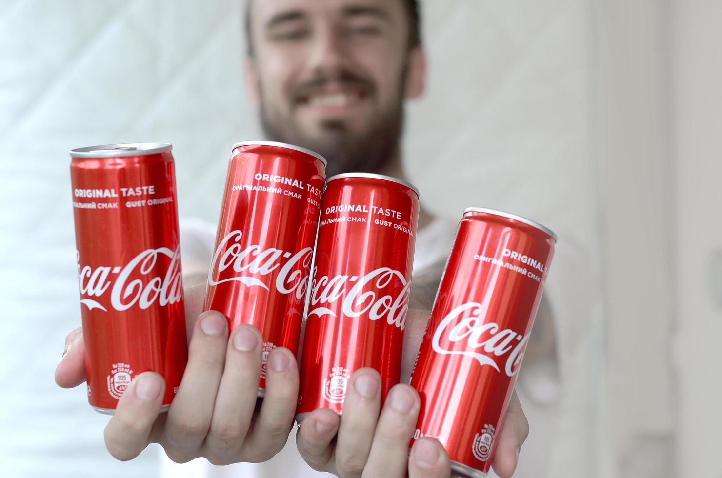 KHARKOV. UKRAINE - MAY 2, 2022 Smiling man holding many non-alcoholic Coca-Cola aluminium tin cans in garage interior photo