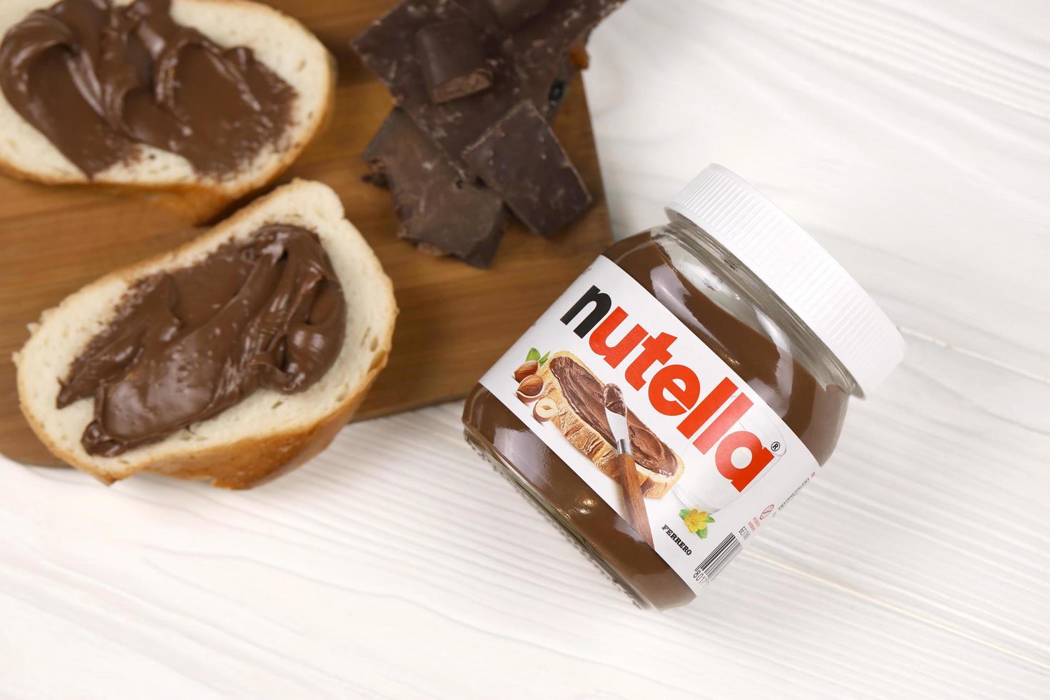 KHARKOV, UKRAINE - MAY 5, 2022 Nutella glass can and spread on freshly baked bread. Nutella is manufactured by Italian company Ferrero first introduced in 1964 photo