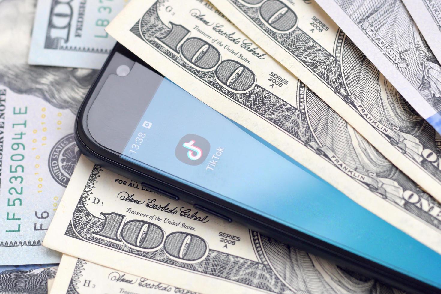 KHARKOV, UKRAINE - MAY 3, 2022 Smartphone screen with Tiktok app and lot of hundred dollar bills. Business and social networking concept photo