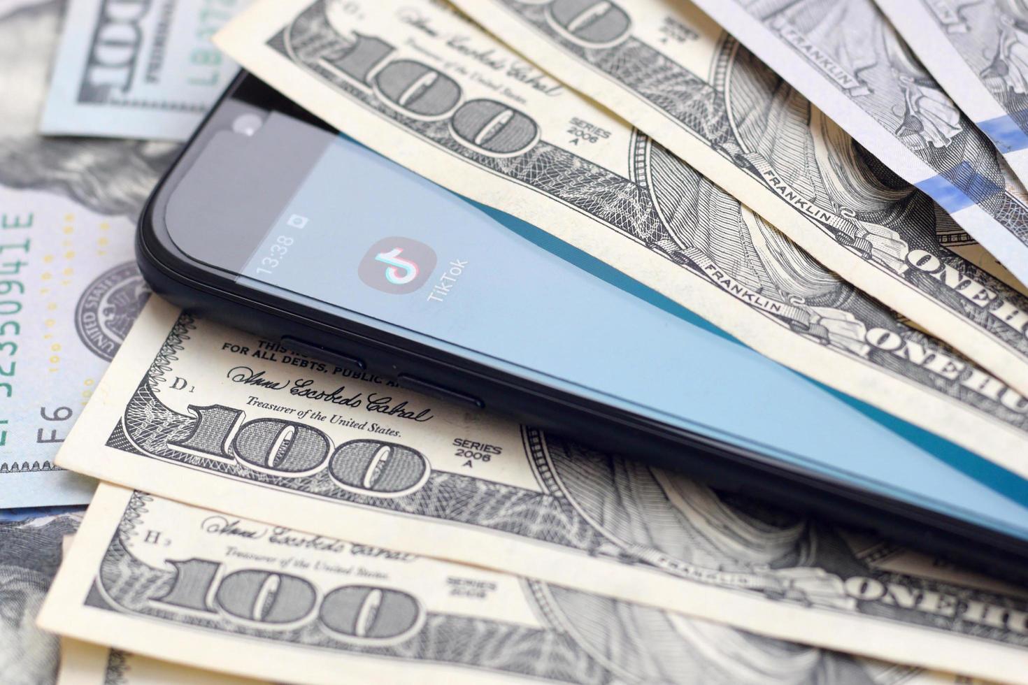 KHARKOV, UKRAINE - MAY 12, 2022 Smartphone screen with Tiktok app and lot of hundred dollar bills. Business and social networking concept photo