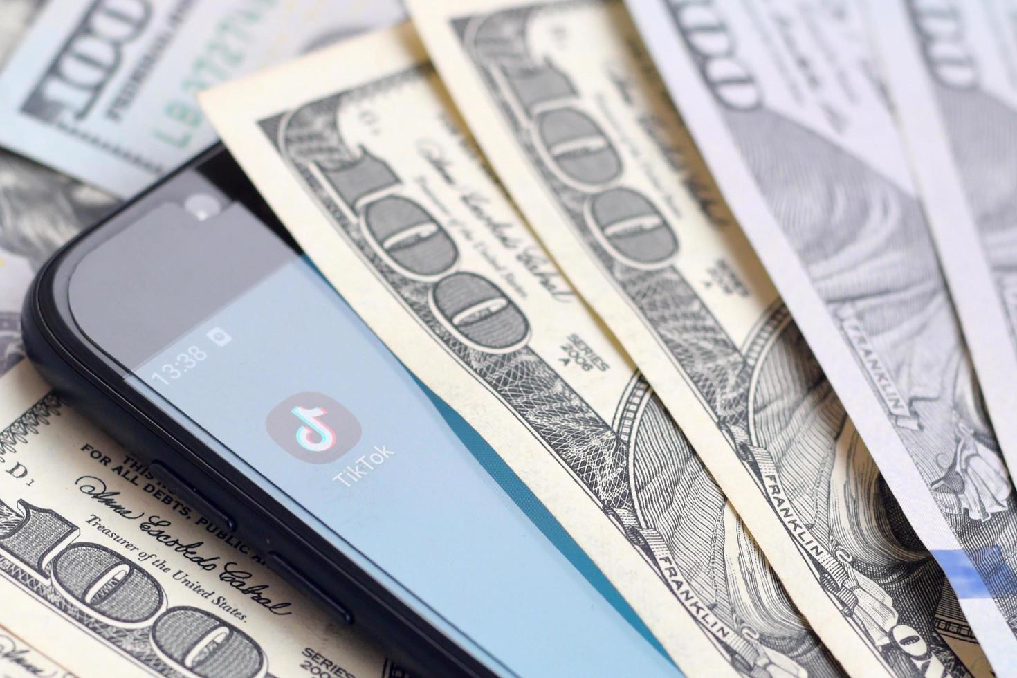 KHARKOV, UKRAINE - MAY 12, 2022 Smartphone screen with Tiktok app and lot of hundred dollar bills. Business and social networking concept photo