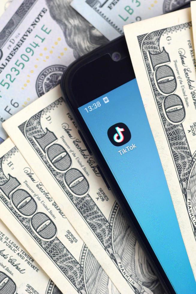 KHARKOV, UKRAINE - MAY 3, 2022 Smartphone screen with Tiktok app and lot of hundred dollar bills. Business and social networking concept photo