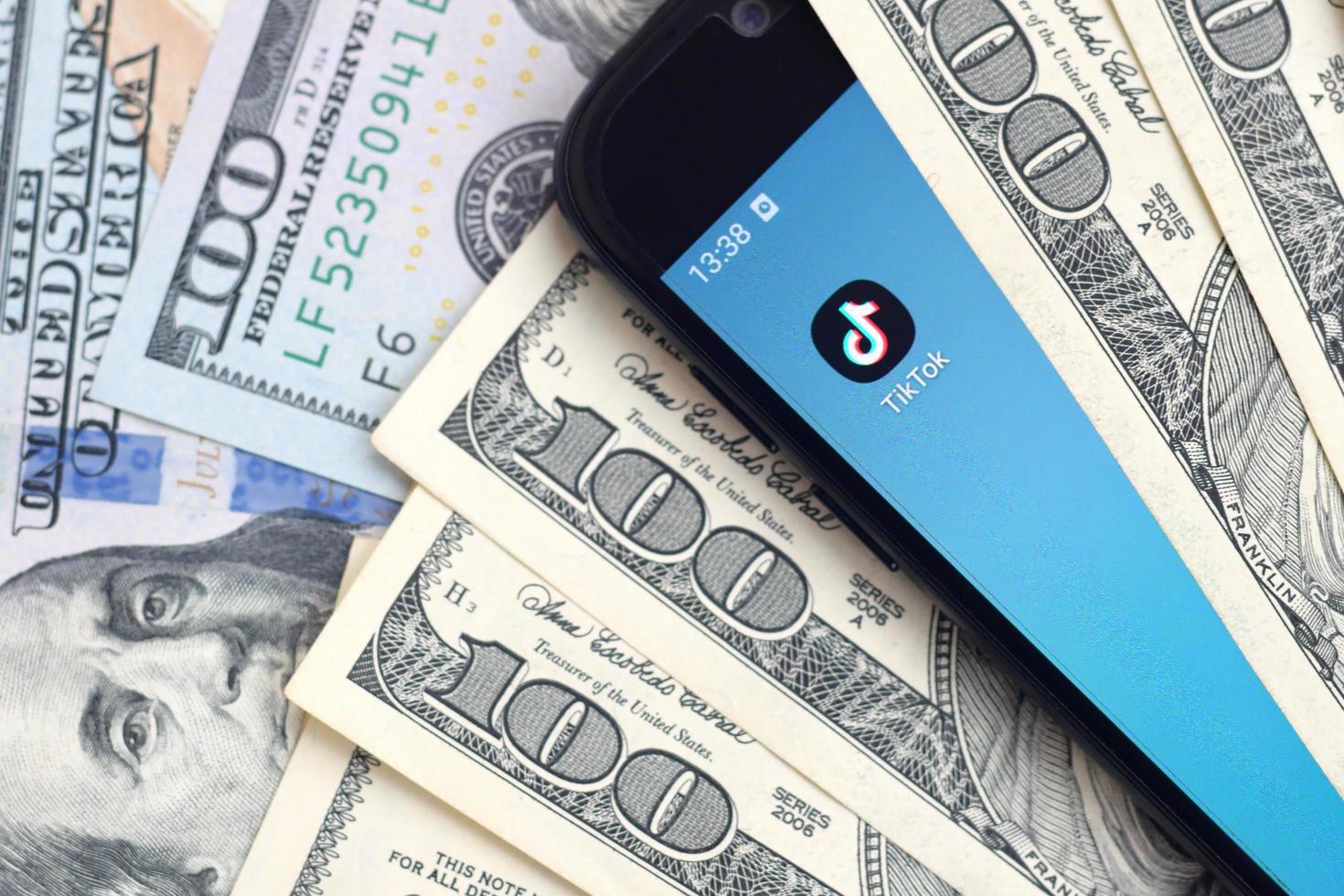 KHARKOV, UKRAINE - MAY 3, 2022 Smartphone screen with Tiktok app and lot of hundred dollar bills. Business and social networking concept photo