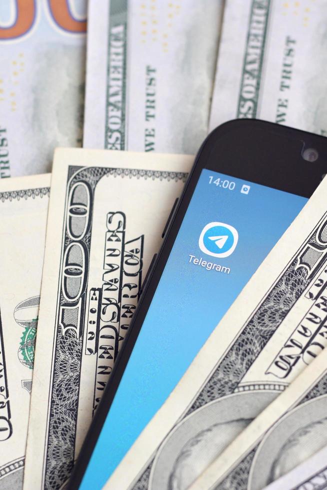 KHARKOV, UKRAINE - MAY 12, 2022 Smartphone screen with Telegram app and lot of hundred dollar bills. Business and social networking concept photo