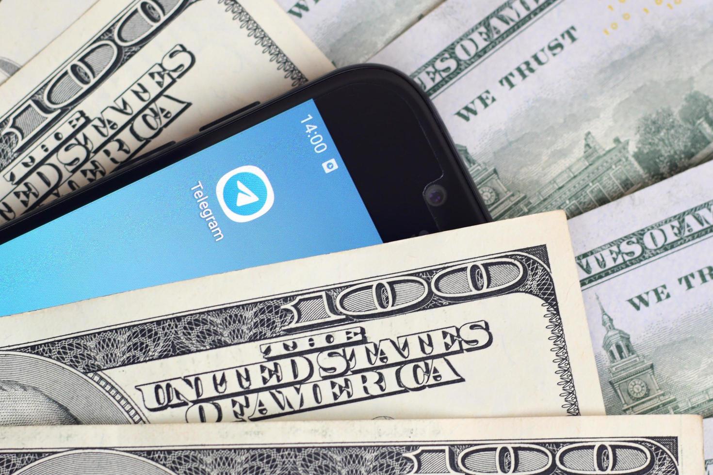 KHARKOV, UKRAINE - MAY 3, 2022 Smartphone screen with Telegram app and lot of hundred dollar bills. Business and social networking concept photo