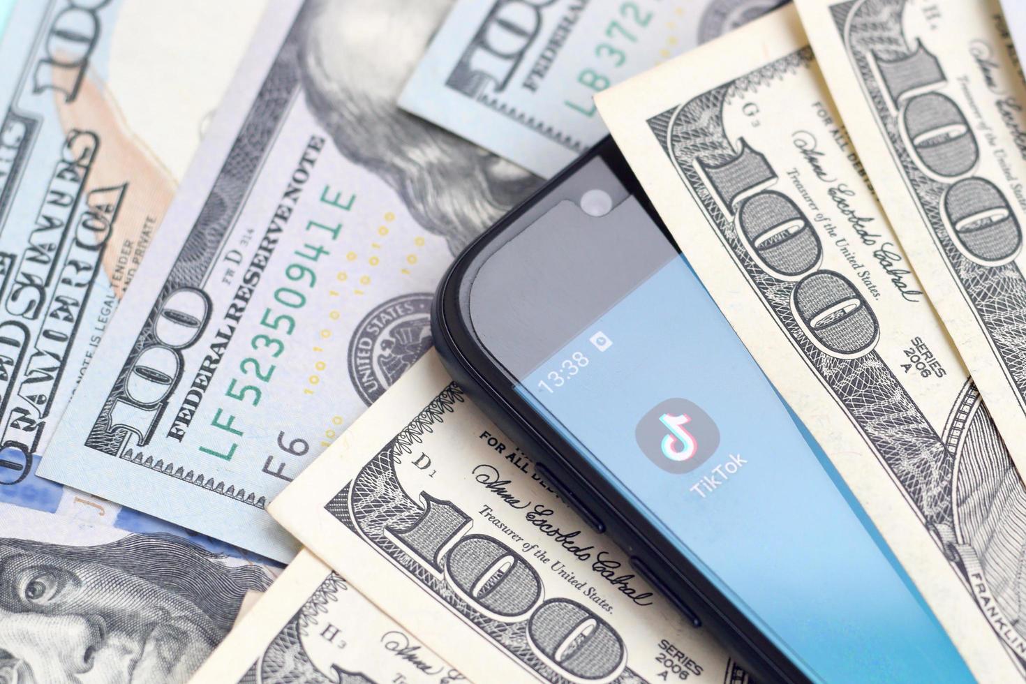 KHARKOV, UKRAINE - MAY 3, 2022 Smartphone screen with Tiktok app and lot of hundred dollar bills. Business and social networking concept photo