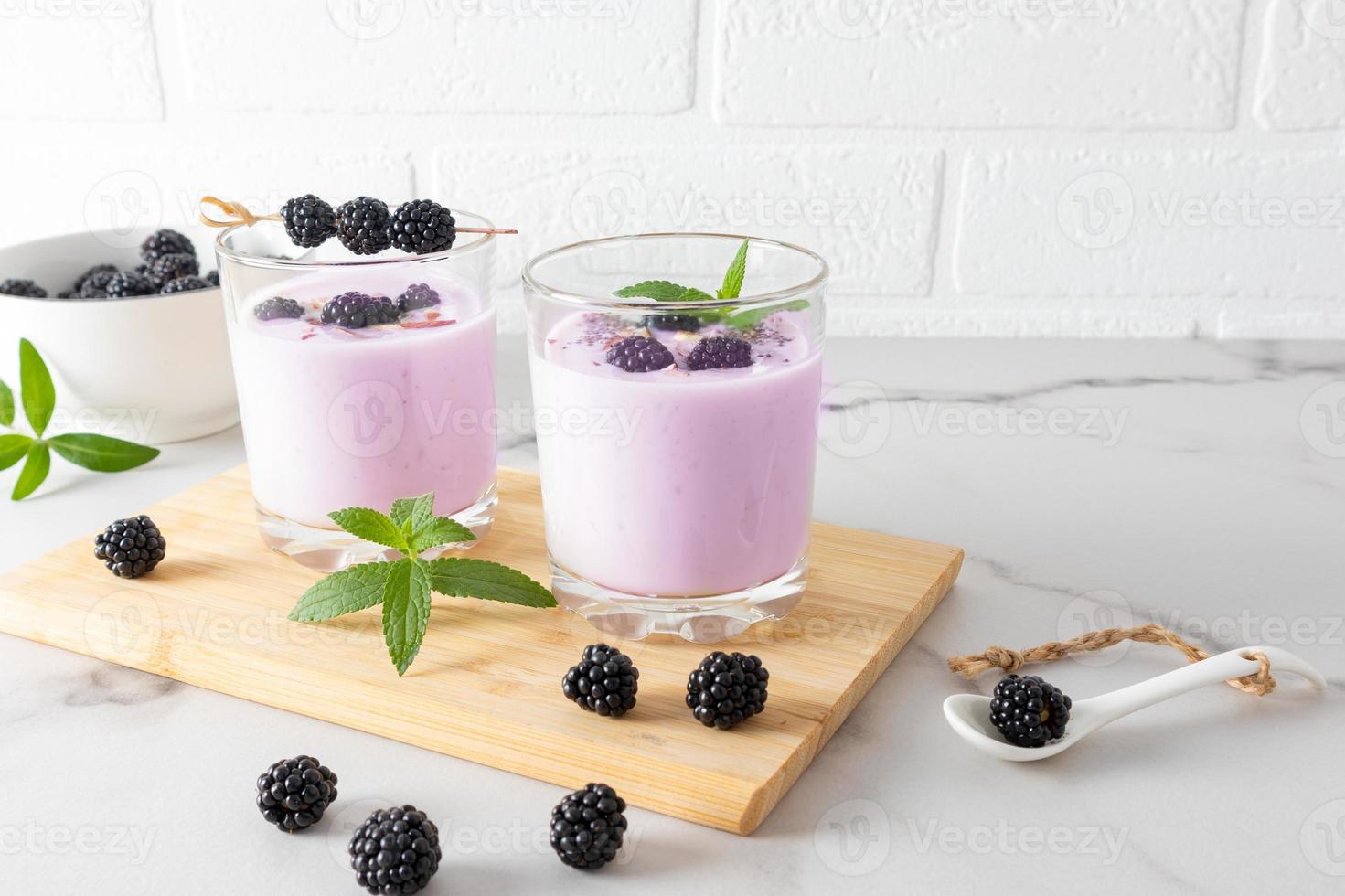 batido de moras en dos vasos sobre una tabla de madera con moras y menta sobre una mesa blanca de mármol contra la pared de ladrillo. alimentación saludable. foto