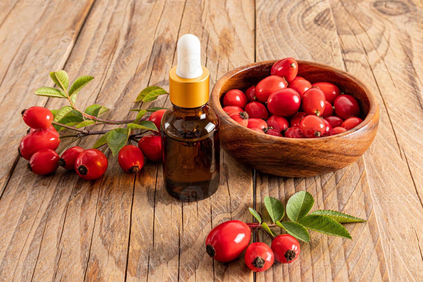 suero para el cuidado de la piel facial a base de aceite de semilla de rosa mosqueta y un bol con frutos frescos de la planta sobre un fondo de madera. biocosméticos naturales. foto