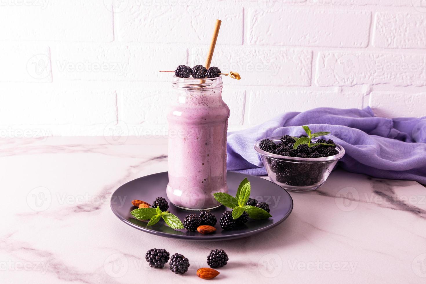 A glass bottle of blackberry smoothie or cocktail stands on a dark ceramic plate on a marble table against a brick wall. healthy eating. diet. photo