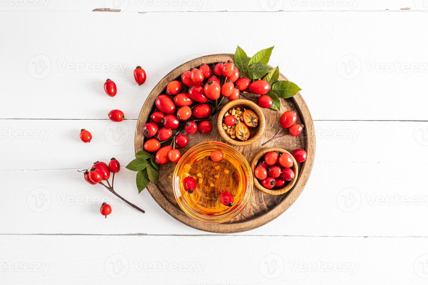 top view of natural rosehip seed oil in a glass bowl on a wooden tray and white wooden table among ripe rose hips. vitamsin C. photo