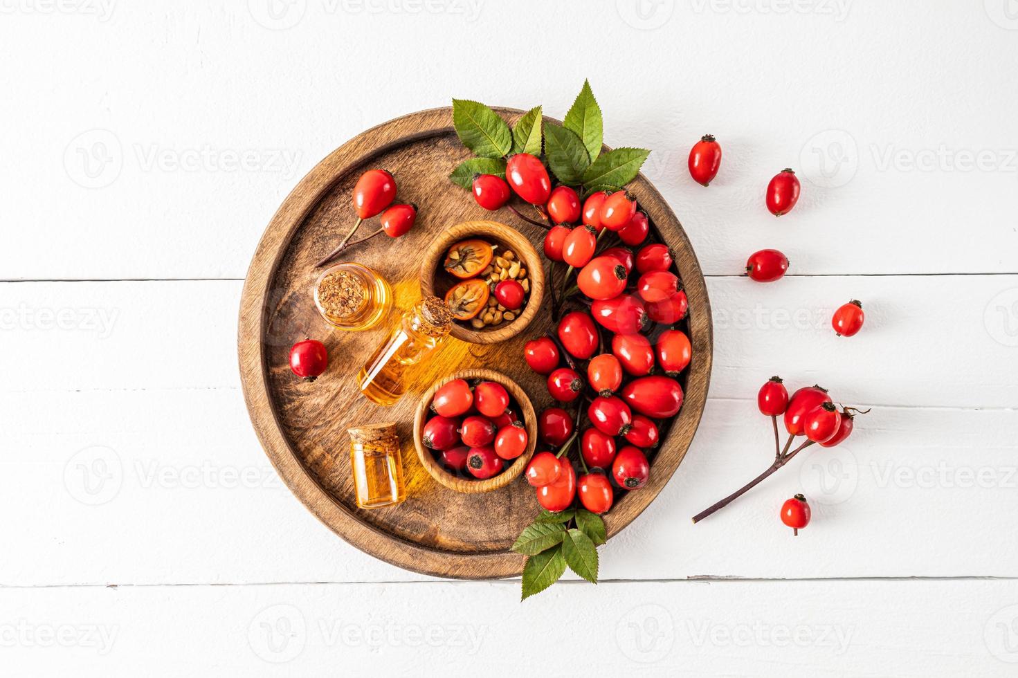 three glass bottles with a cork with organic rosehip seed oil on a wooden round tray among ripe berries. top view. white wooden background. photo