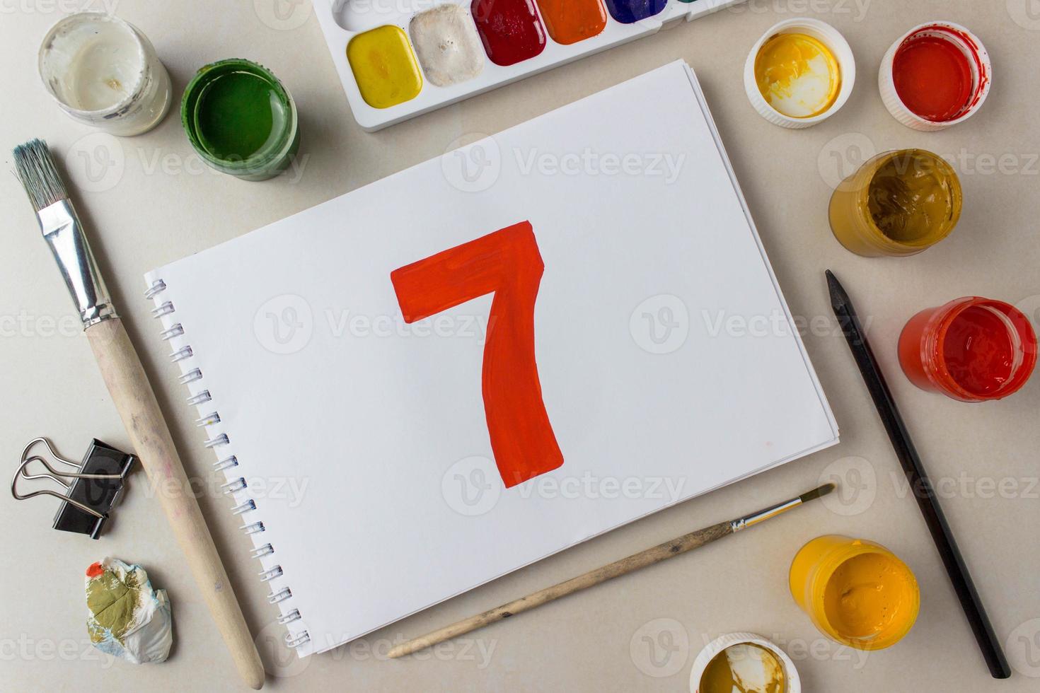 Red number seven drawing on a white album sheet. To indicate the place, price, etc. Nearby are brushes, jars of paint gouache, watercolor, etc photo