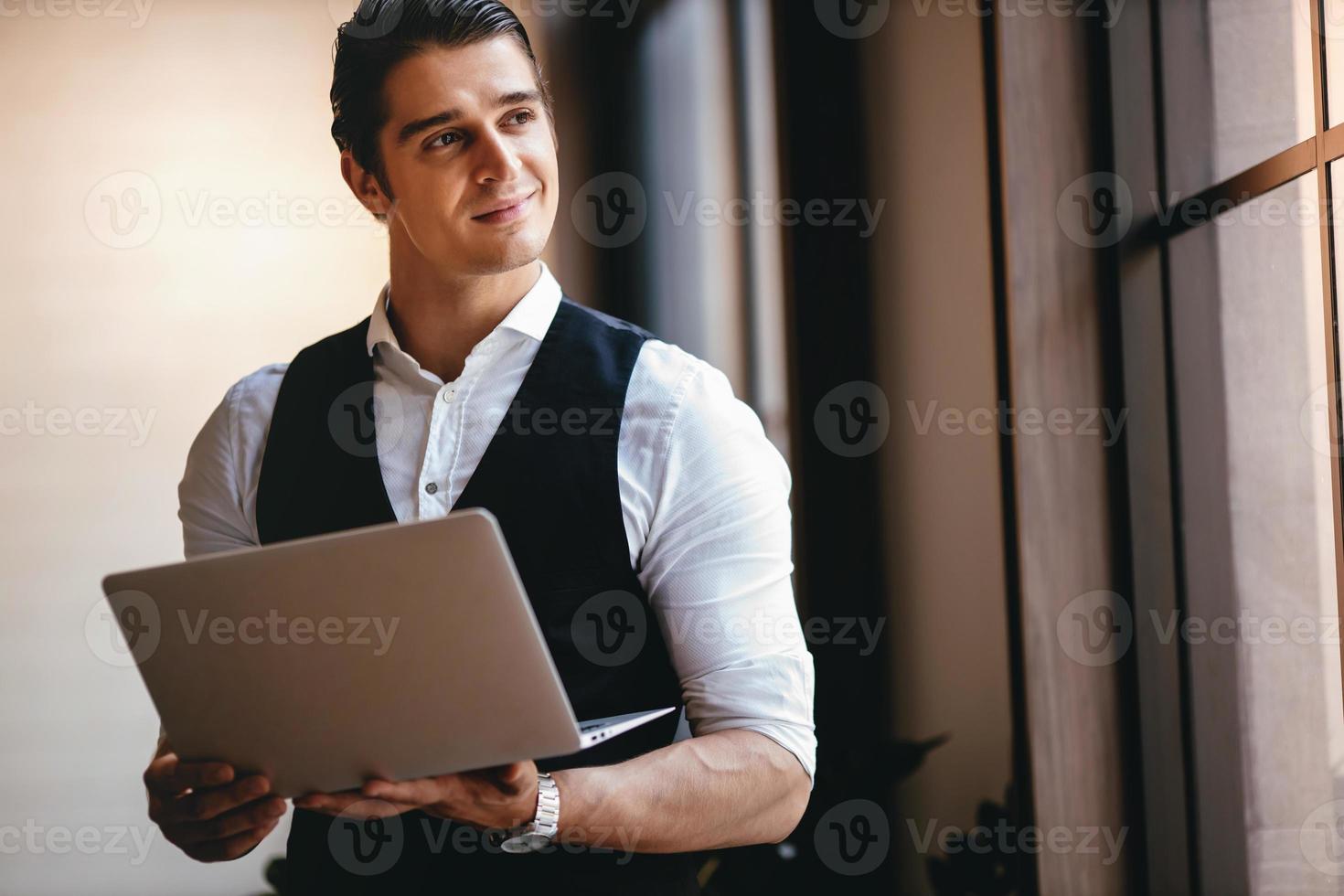 a Young Caucasian Businessman Working on Computer Laptop in the Modern Workplace. Smiling Businessman Standing by the Window and Looking outside photo