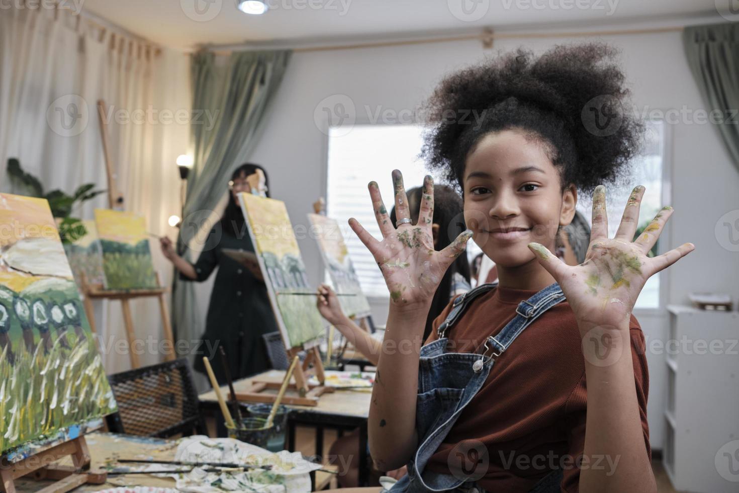 African American girl shows her hand messed up with acrylic colors, smiles and looks at camera, fun learns with student kids in art studio class, creative painting with skills in school education. photo