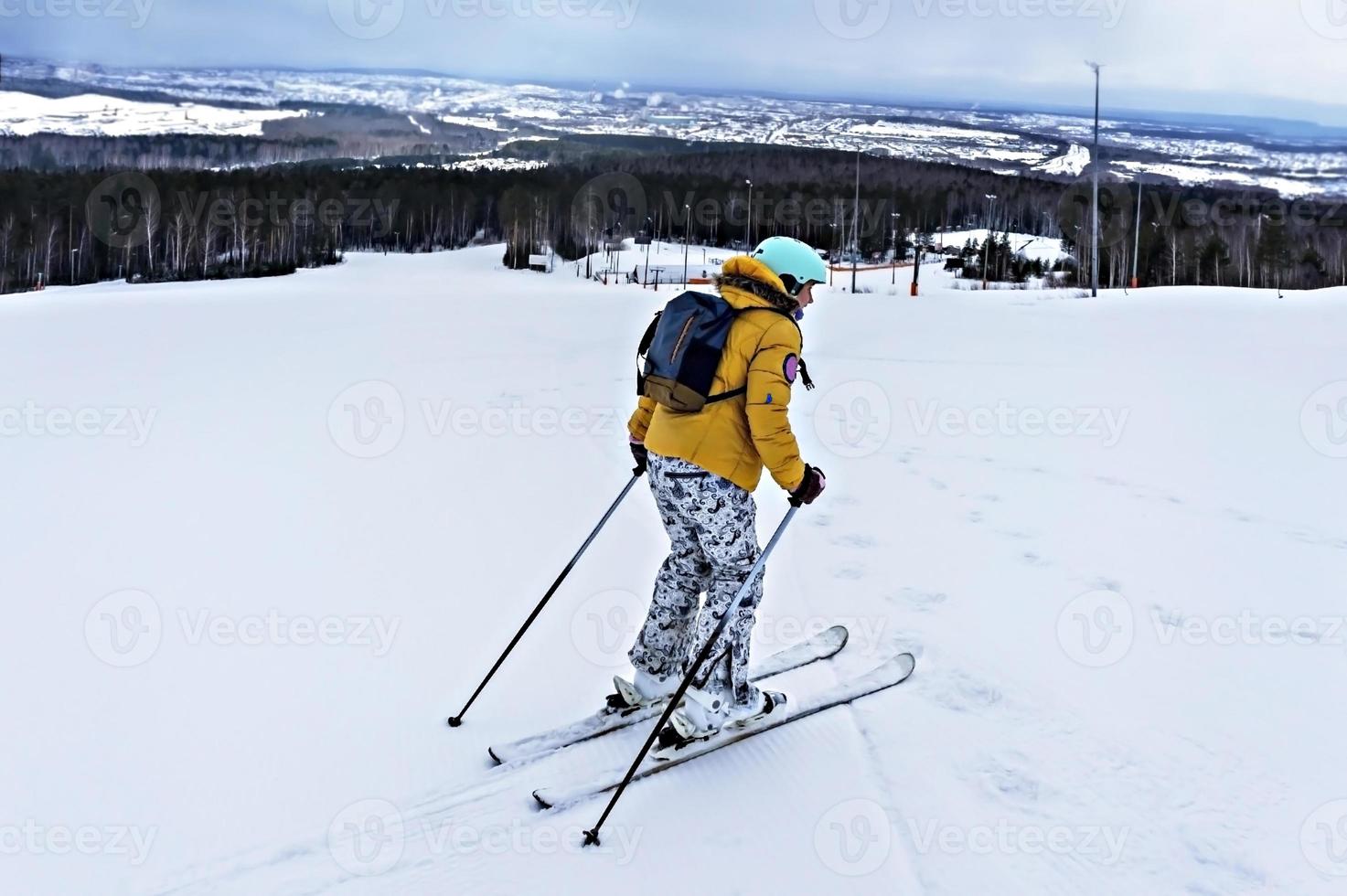 mujer joven con chaqueta amarilla y casco de esquí esquiando en una ladera de montaña, deportes de invierno, actividad de esquí alpino al aire libre, estilo de vida saludable foto