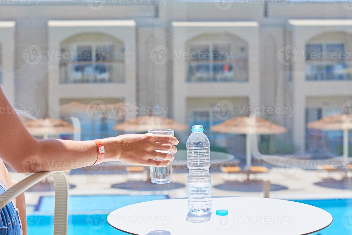 a woman hand hold glass of water at hotel balcony against swimming pool photo