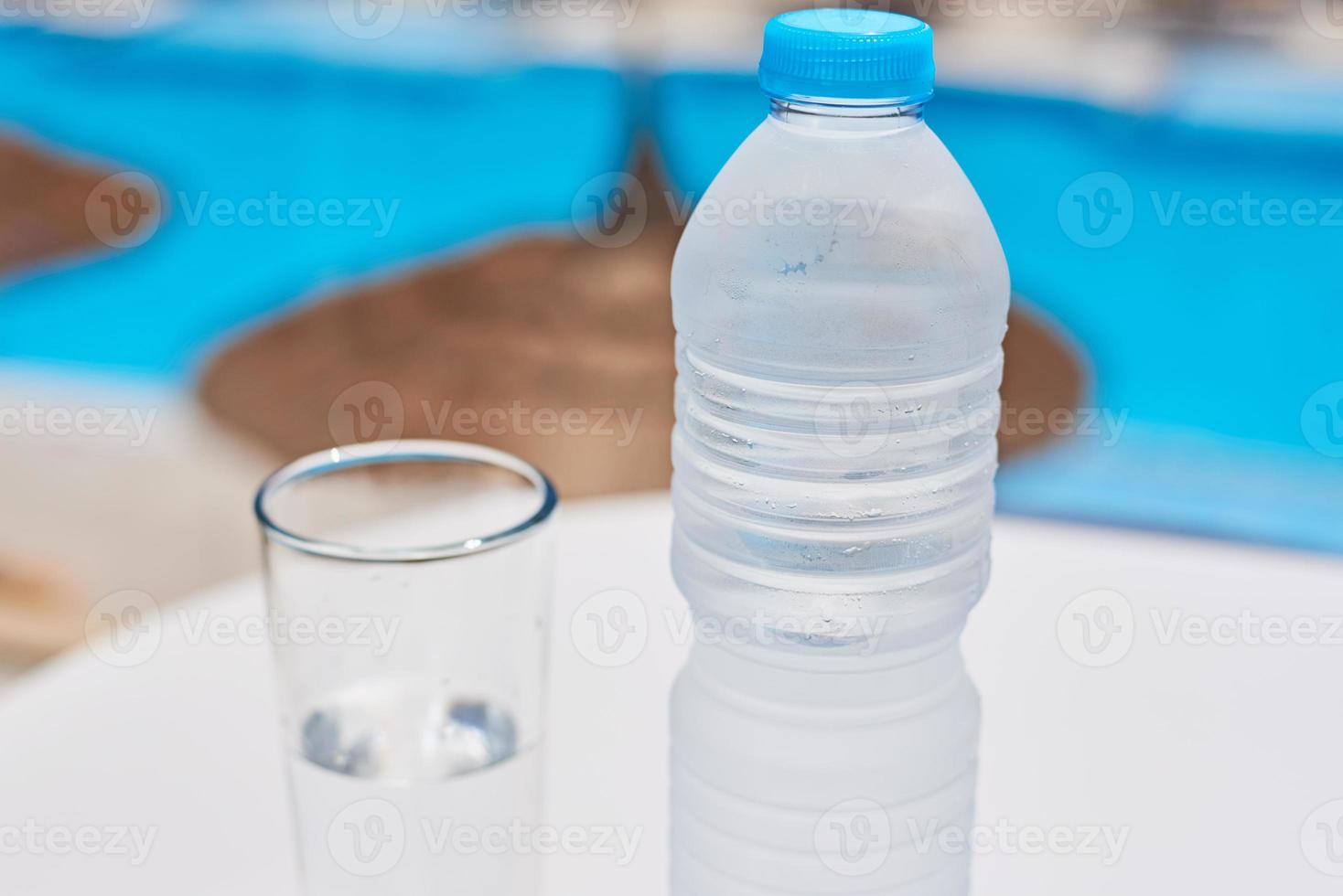 Glass and bottle with a water on white table agains swimming pool background photo