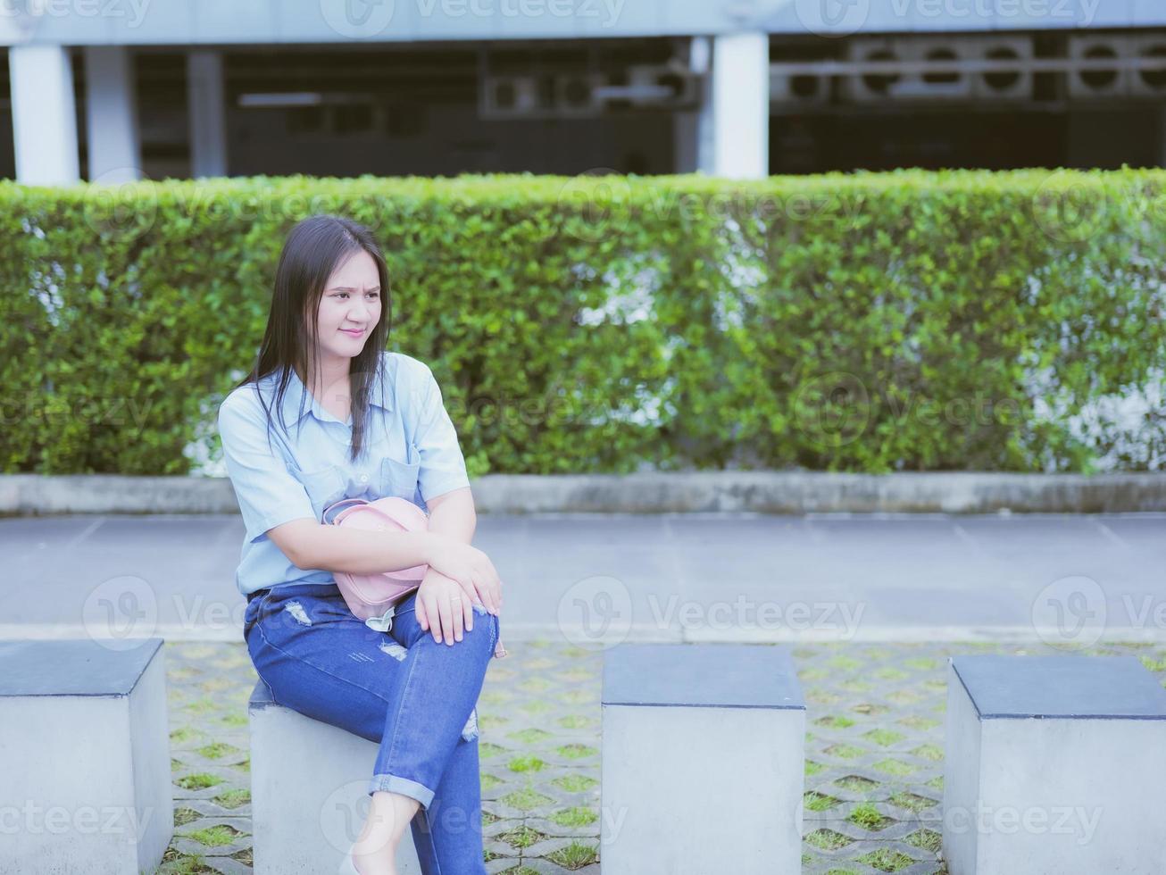 Take a portrait of an Asian woman with black hair in a blue shirt and jeans. sit on the ninth in the park and has a background of buildings and bushes. photo