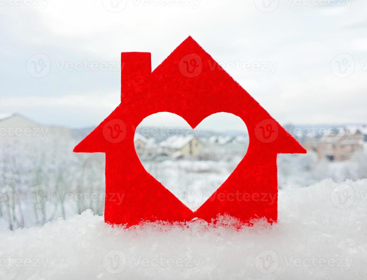 Small house cut out of red felt is standing in the snow. Heart-shaped hole is cut instead of a window. Blurred background of cottage village in winter. Valentine's day concept. photo