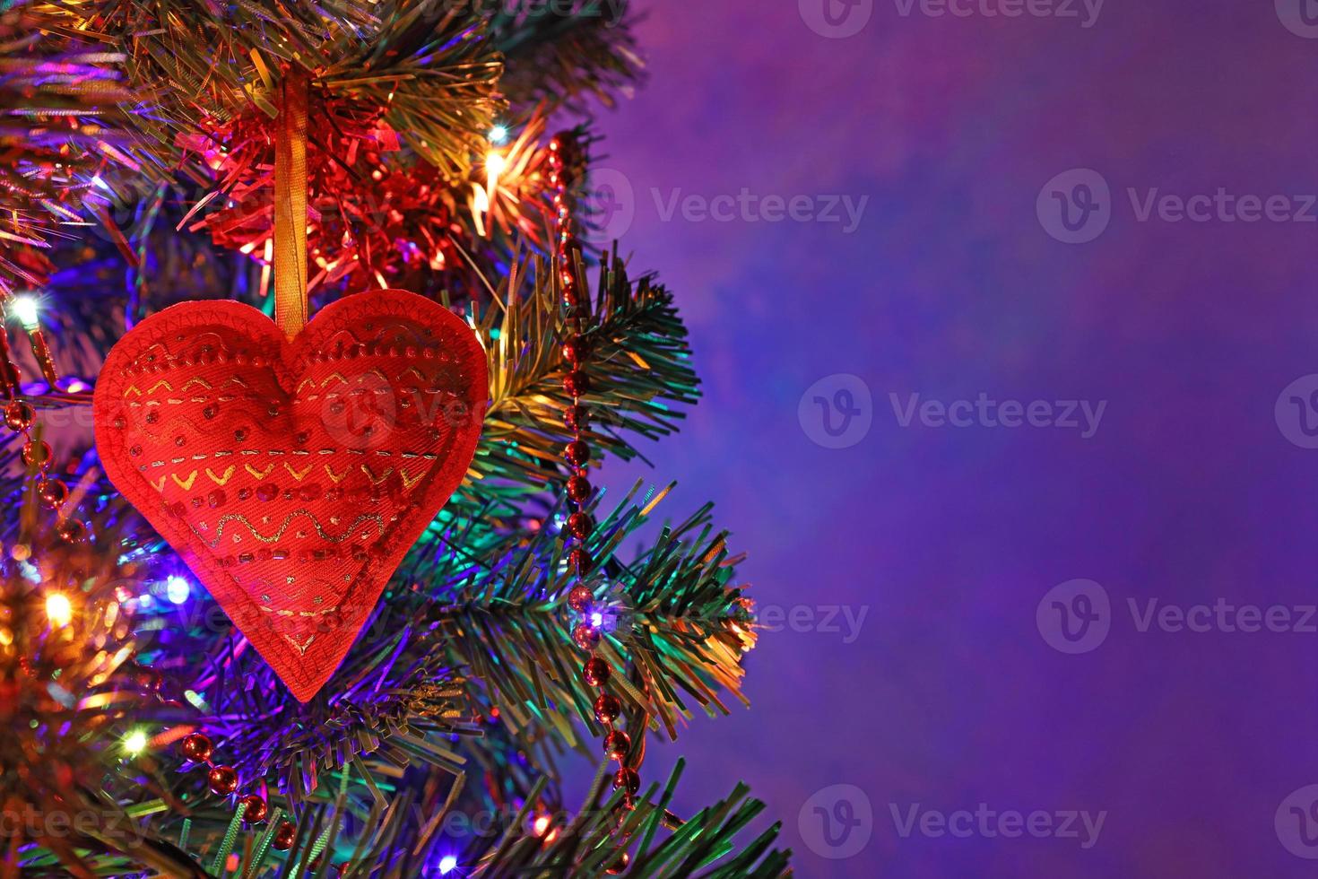 Red heart Christmas handmade decoration with ornament on fir tree at holiday night, colorful bokeh garland, red beads, blurred dark purple background. Xmas greeting card with copy space. photo