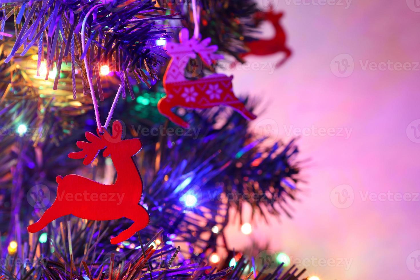 Christmas greeting card with three red santa's reindeers, one close up. Traditional wooden decorations with ornament on Christmas fir tree. Colorful bokeh garland, blurred background, purple backlight photo