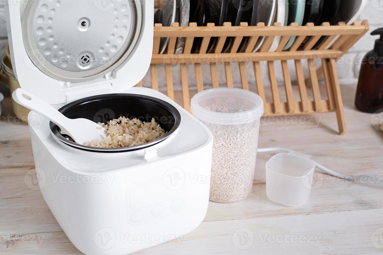 Opened electric rice cooker with cooked steaming brown rice on wooden counter-top in the kitchen photo