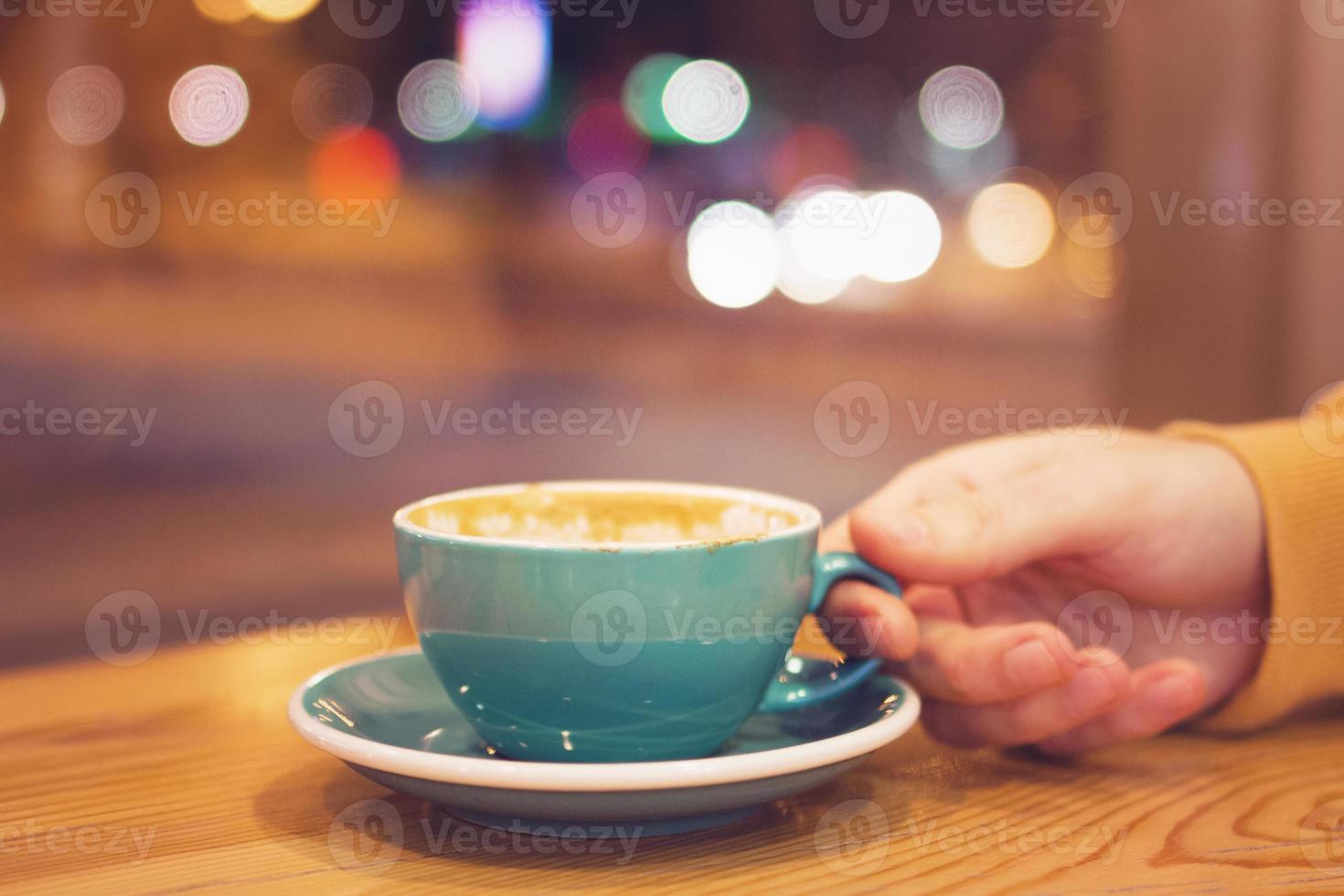 mano de hombre con una taza de café en un café sobre un fondo de luces de la ciudad detrás de la ventana. foto tonificada.