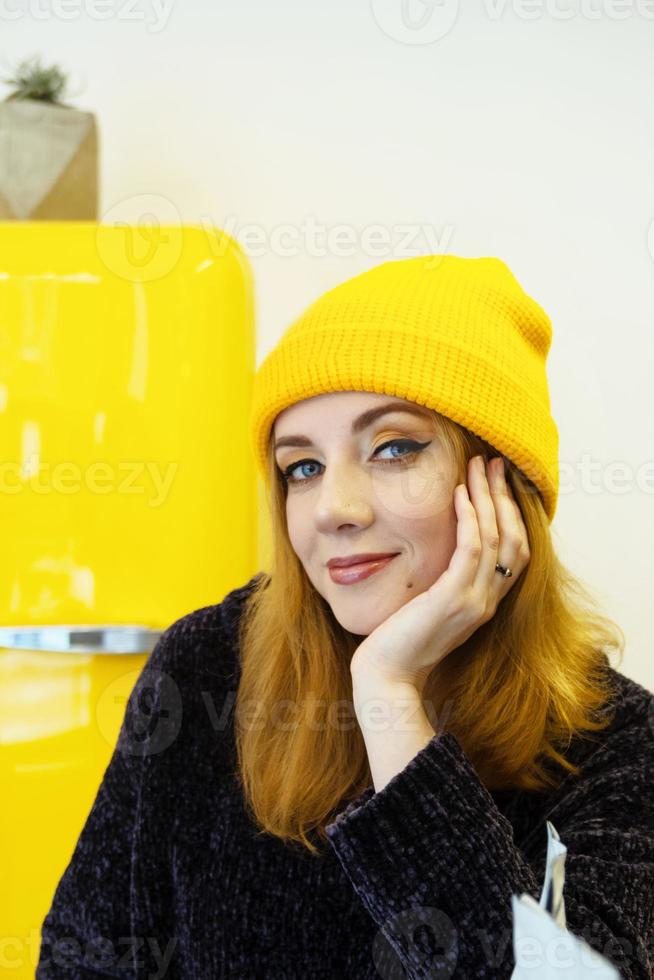 Portrait of young woman with blond hair in a yellow knitting hat is sitting in a cafe near to yellow fridge. photo