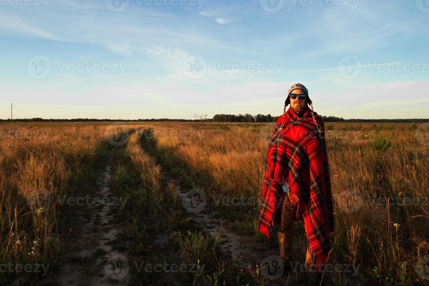 Young happy smiling man in the knitted cap, sunglasses and blanket is meeting of sunrise on the background of a field and blue sky. photo