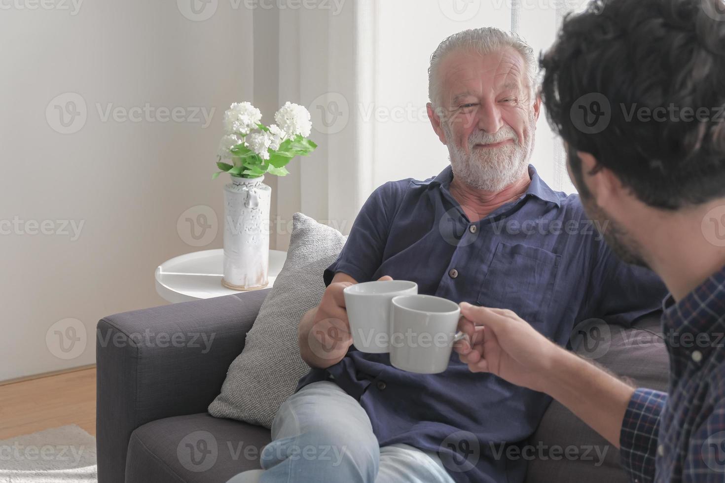 Family relaxation concept. Two of old father and son are clinking coffee cups together with happy smiling face during sitting at living room cozy warmth house on holiday, rear view and copy space. photo