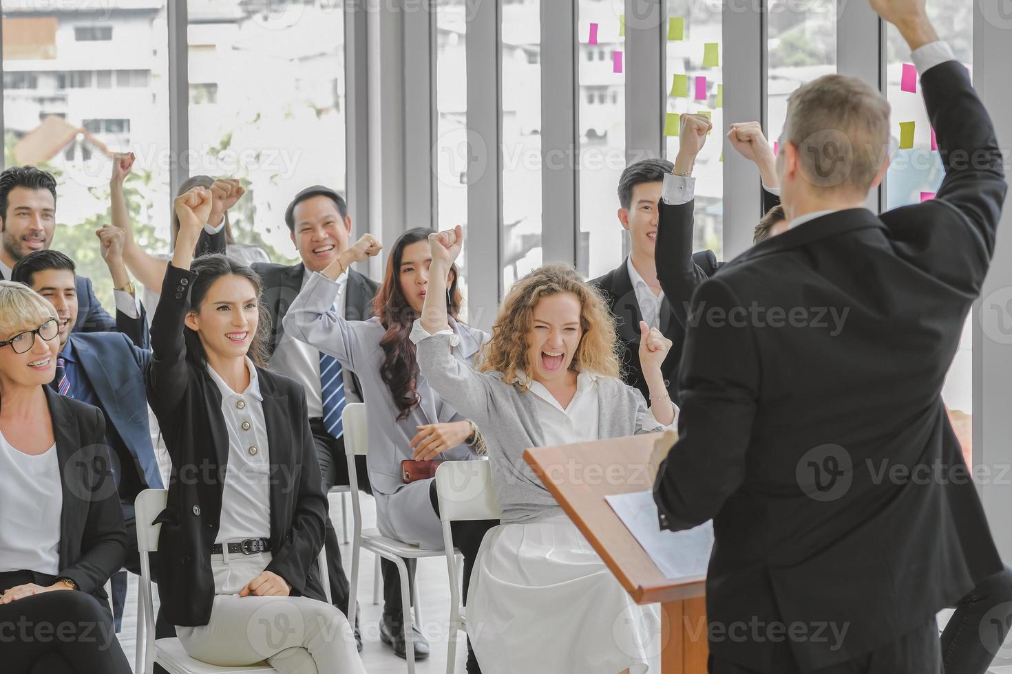 feliz audiencia multiétnica de negocios en la sala de conferencias levantando los puños junto con caras sonrientes siguen al orador público masculino en el podio en el evento del seminario corporativo, vista trasera. foto