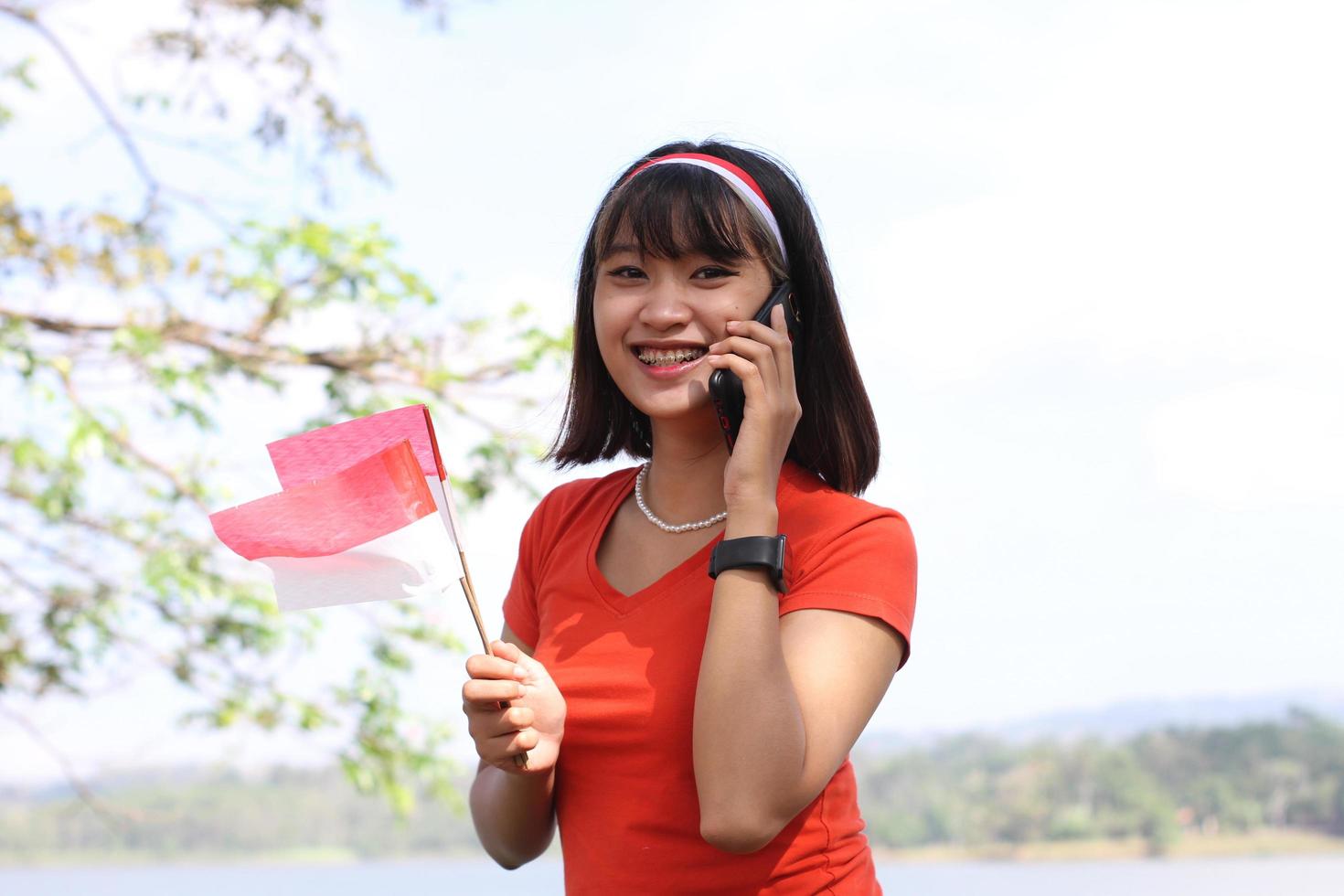 hermosa joven asiática que lleva la bandera indonesia con una cara alegre y llama a su amigo foto