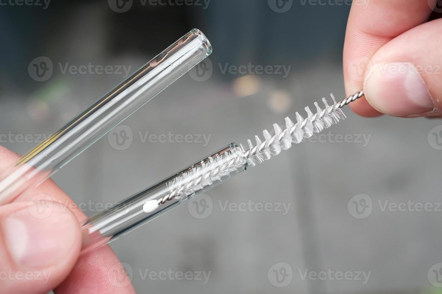 Hands holding reusable glass straws for cocktails and cleaning them with a special brush on a blurred gray background. Ecological concept. photo