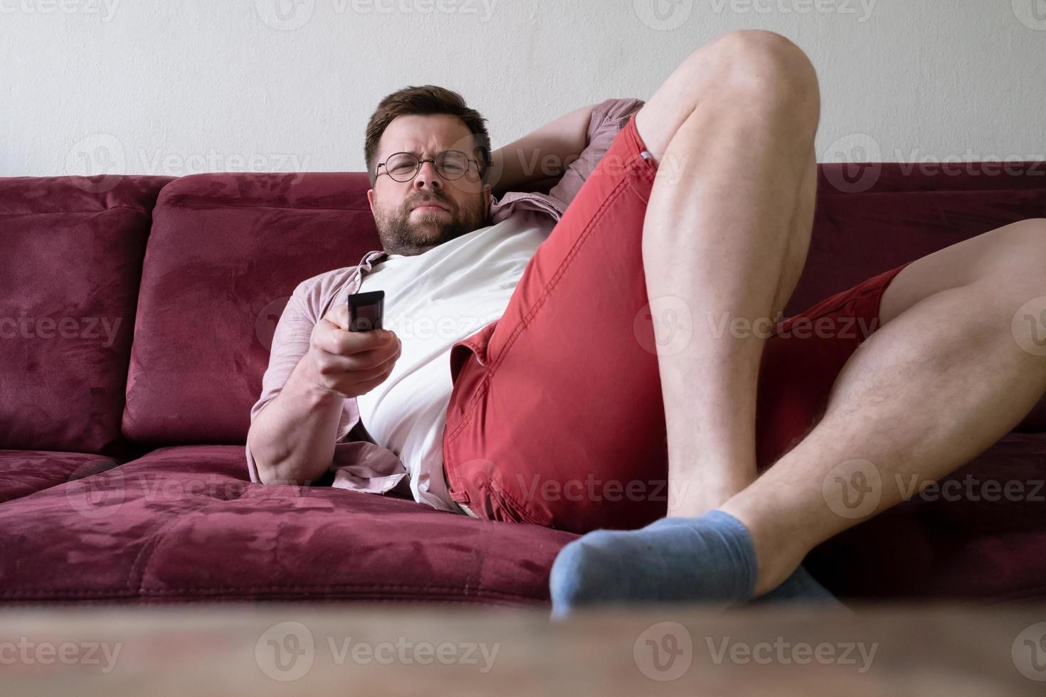 Tired, frowning man holds a TV remote while lying on a sofa and watching movies. Concept of lazy rest. photo