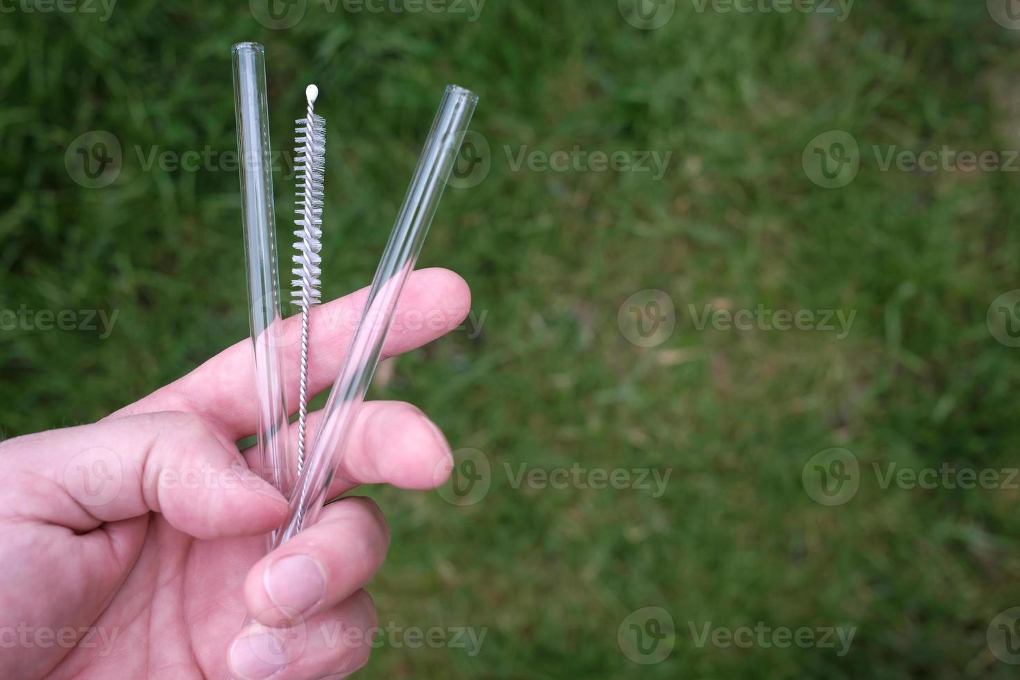 Hands hold reusable glass straws for cocktails and a special cleaning brush, on a background of greenery. Ecological concept. photo