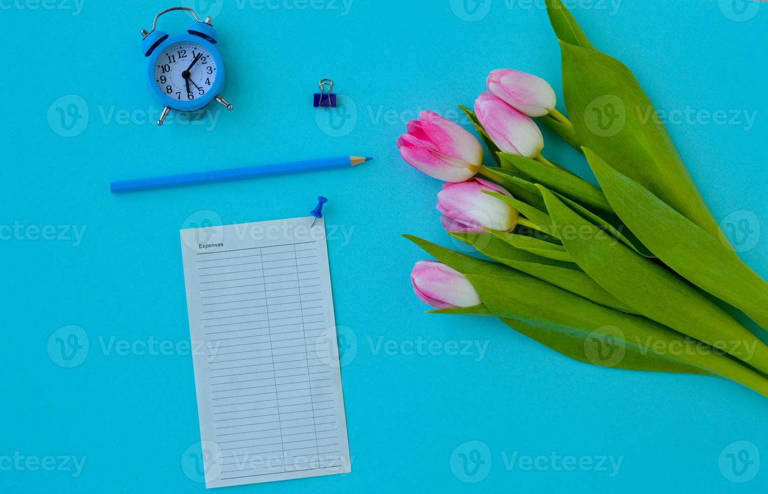 Flat lay woman home office desk. Workspace with pink tulips bouquet, alarm clock, pencil and a piece of paper for notes on blue background. Top view feminine background. photo