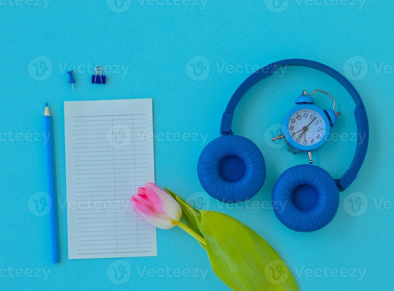 Flat lay home office desk. Workspace with pink tulip, headphones, alarm clock, pencil and a piece of paper for notes on blue background. Top view feminine background. photo