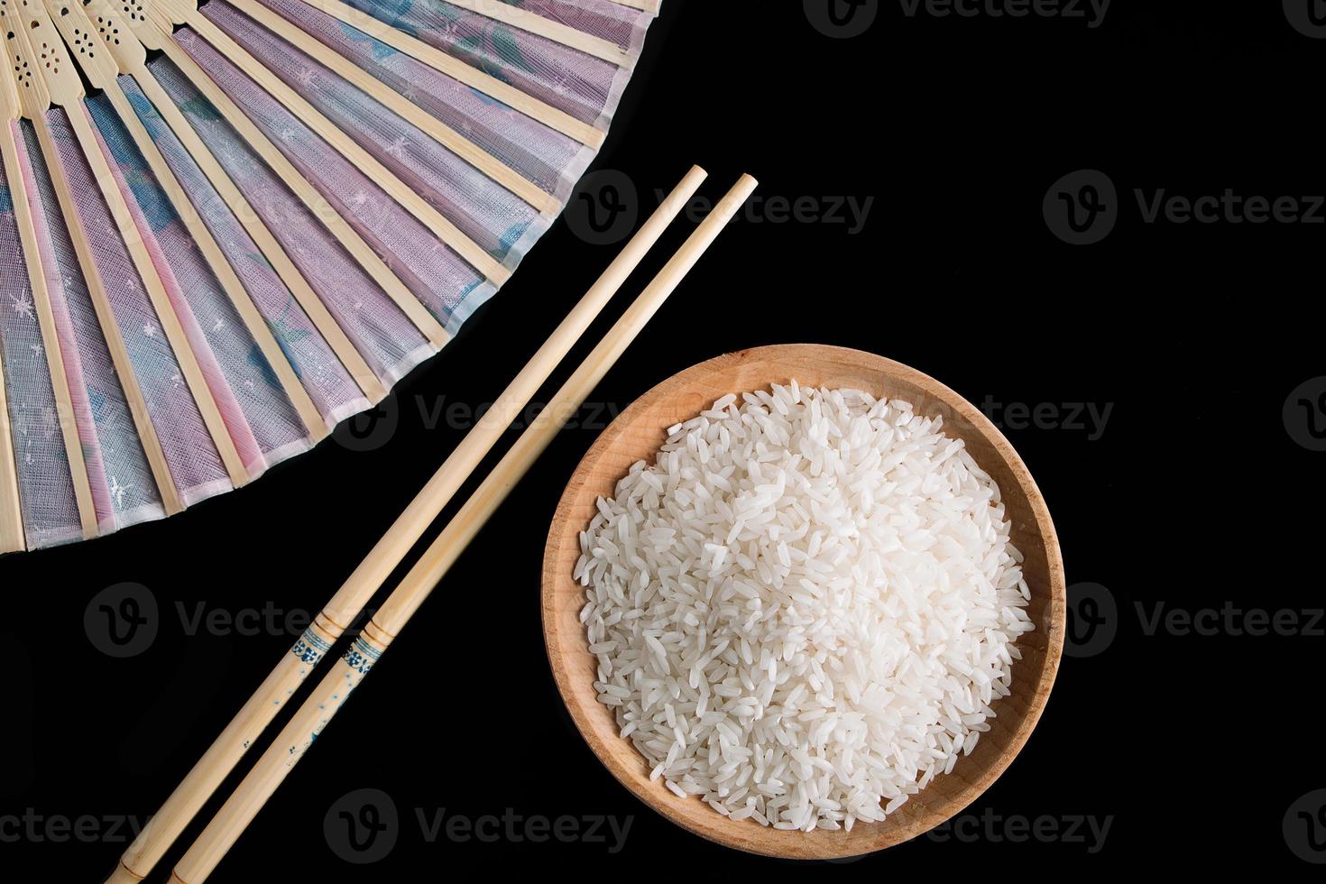 Raw white rice, hand fan and chopsticks on black background. Long uncooked rice in a wooden plate. photo