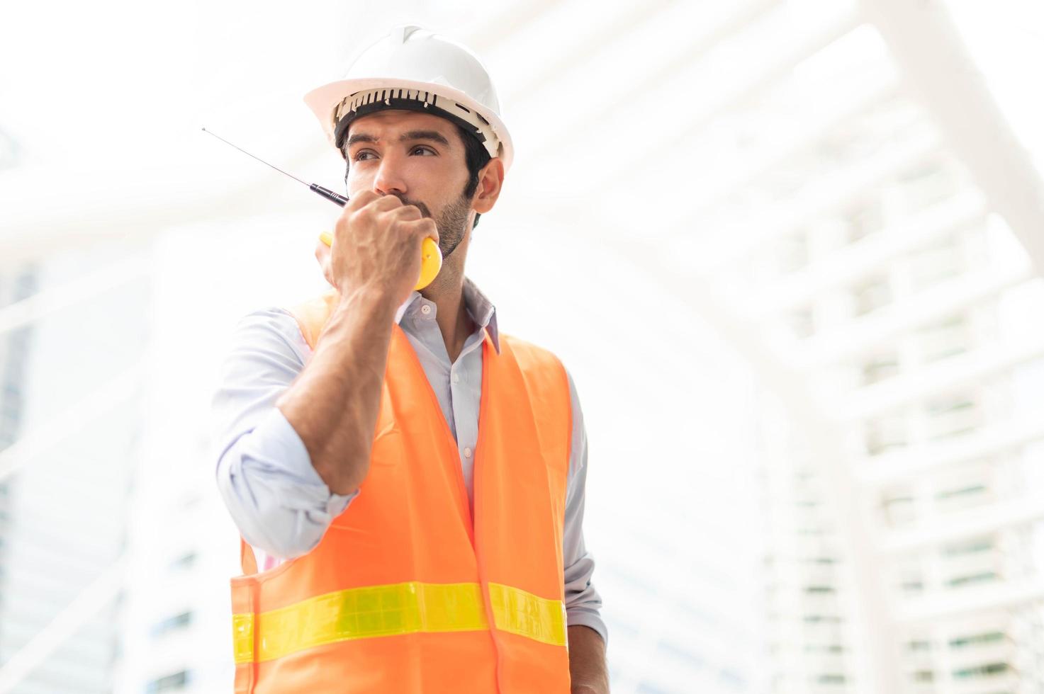 el ingeniero caucásico usa un walkie-talkie para hablar, usa chaleco naranja y un gran sombrero duro en el sitio de trabajo del centro de la ciudad. foto