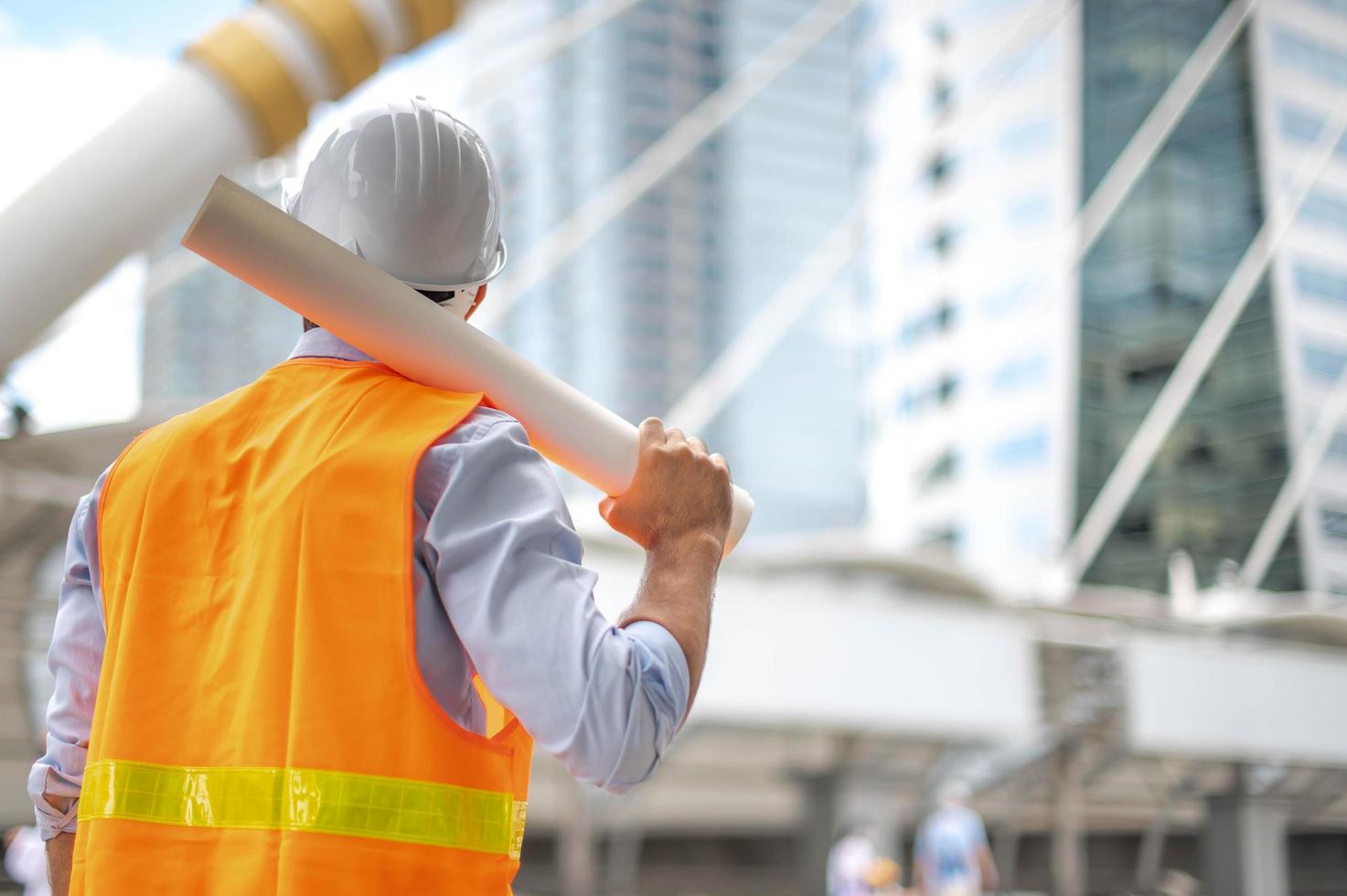 joven caucásico sosteniendo un papel grande, un tipo con camisa azul claro y jeans con chaleco naranja y casco blanco para seguridad en el área de construcción. foto