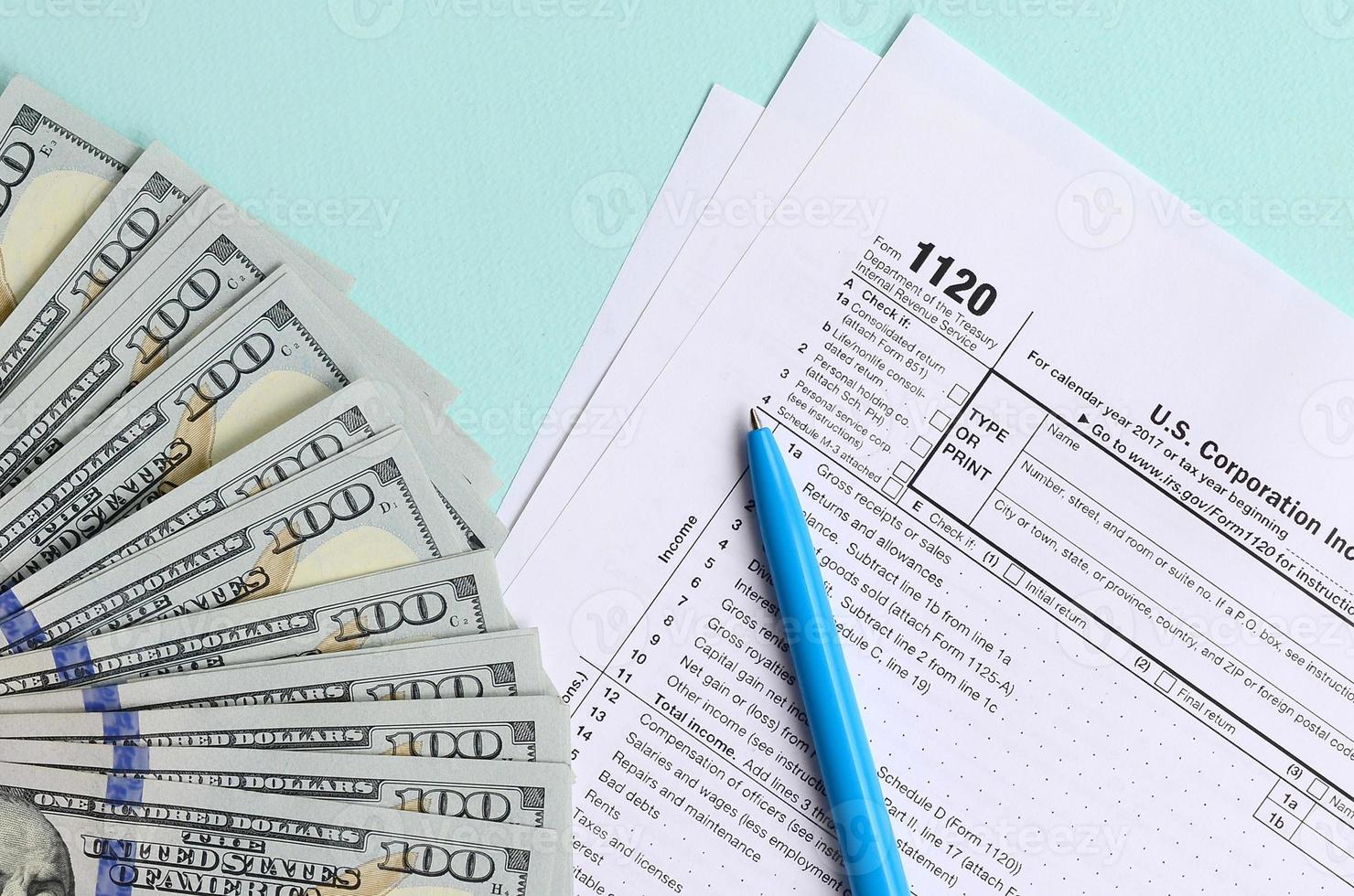 1120 tax form lies near hundred dollar bills and blue pen on a light blue background. US Corporation income tax return photo