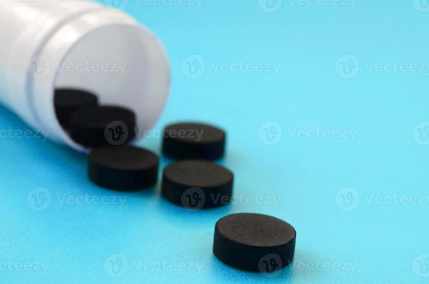 varias tabletas negras caen del frasco de plástico en la superficie azul. imagen de fondo sobre temas médicos y farmacéuticos. carbón activado foto
