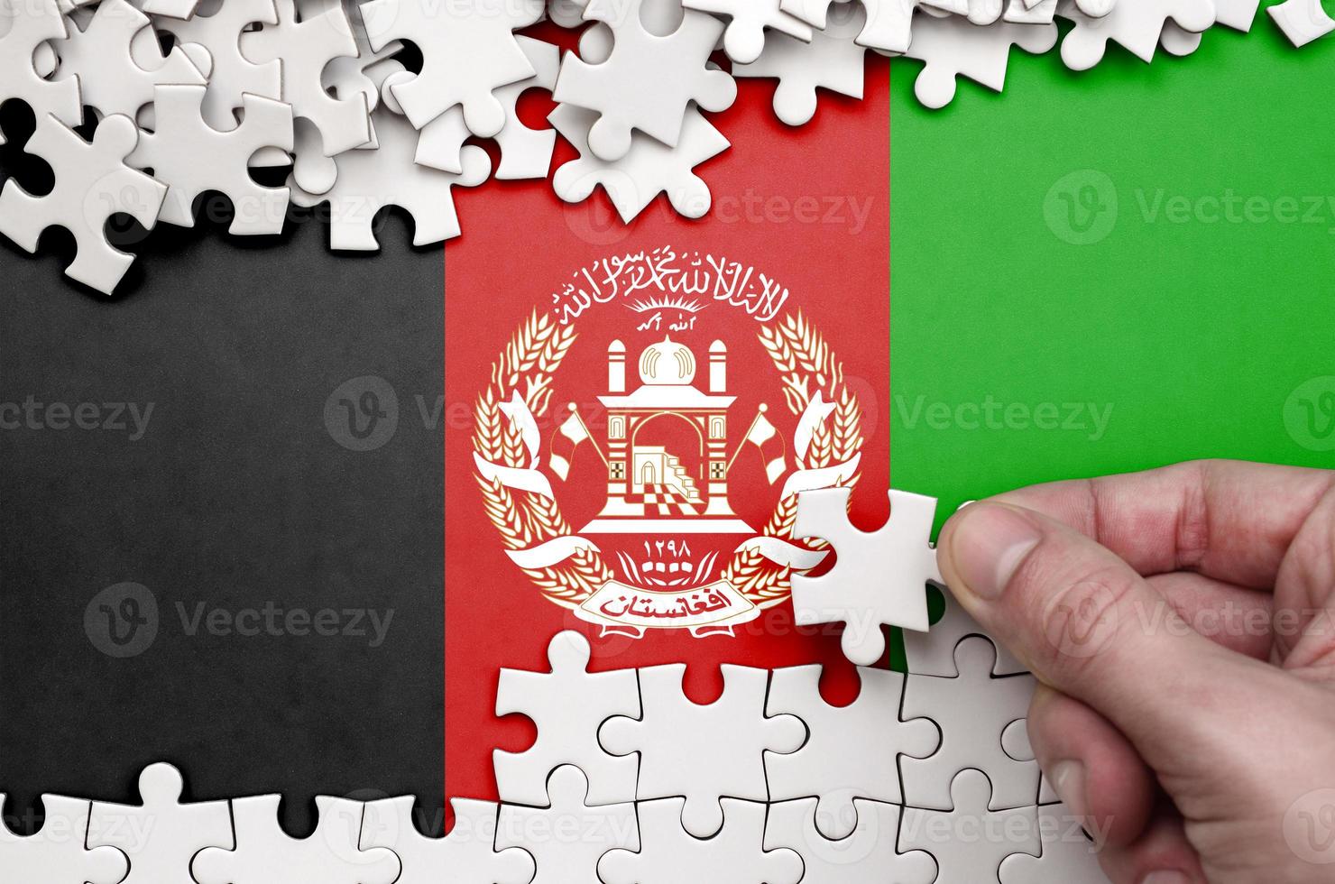 Afghanistan flag is depicted on a table on which the human hand folds a puzzle of white color photo